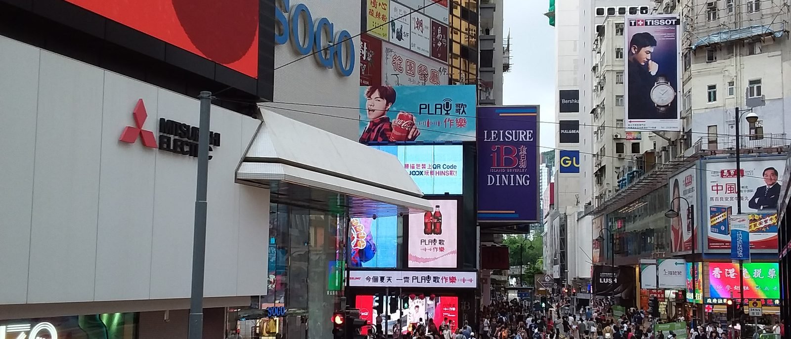 A busy night-time street corner in Jardine's Bazaar, Causeway Bay, a bank branch and neon shop signs above pedestrians crossing near a minibus stop.