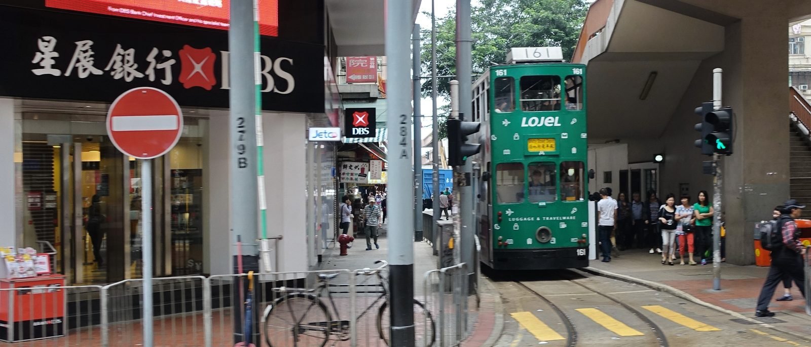 A green tram nearing the North Point terminus, passing a DBS bank branch as pedestrians wait to cross at the signal.